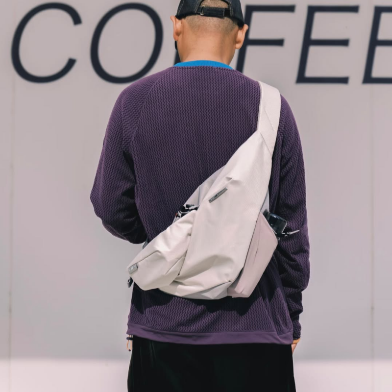 Back view of a male model wearing purple textured long sleeve top and backward black cap, carrying NIID Radiant R2 gray shadow grid modular sling bag crossbody, light wall with large 