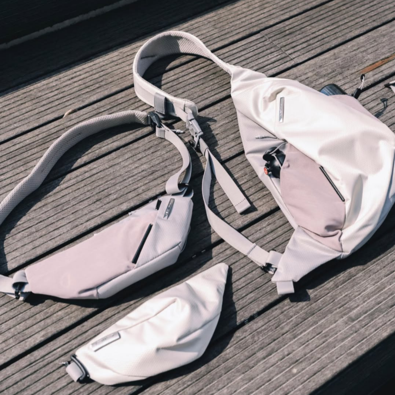 Flat lay top-down shot of complete NIID Radiant R2 modular sling bag system, including main crossbody bag, attachable wing pocket and mini tech sling pouch, light gray shadow grid fabric, laid out on weathered wooden outdoor deck, natural soft daylight, minimalist product display, no watermarks
