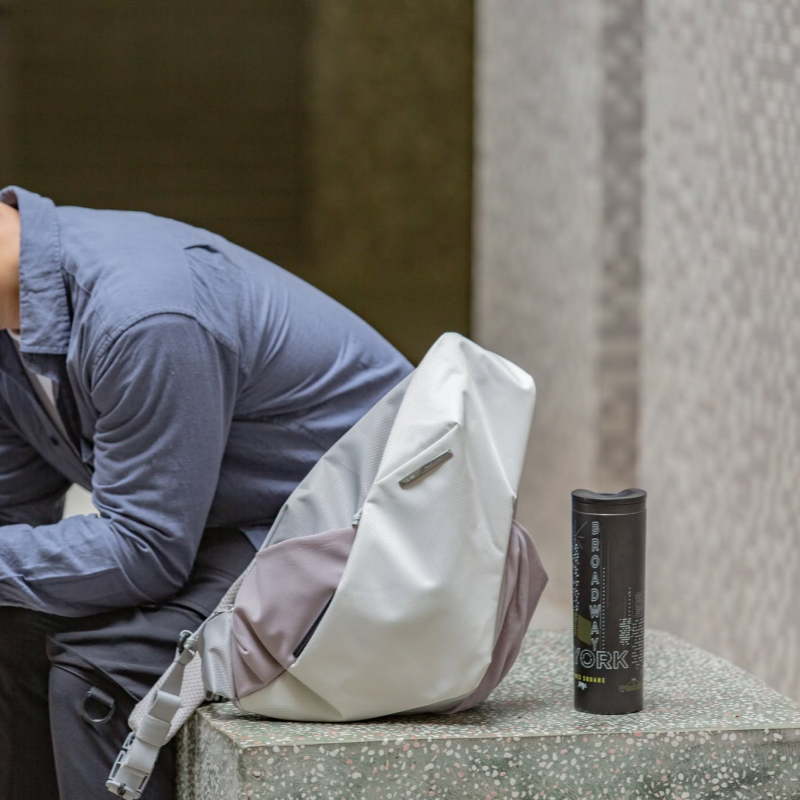 Lifestyle shot of NIID Radiant R2 gray shadow grid modular crossbody sling bag placed on speckled stone bench with black insulated water tumbler beside it, man in light blue casual shirt sitting nearby, minimalist urban neutral tone scene, waterproof functional everyday chest bag, clean no watermark
