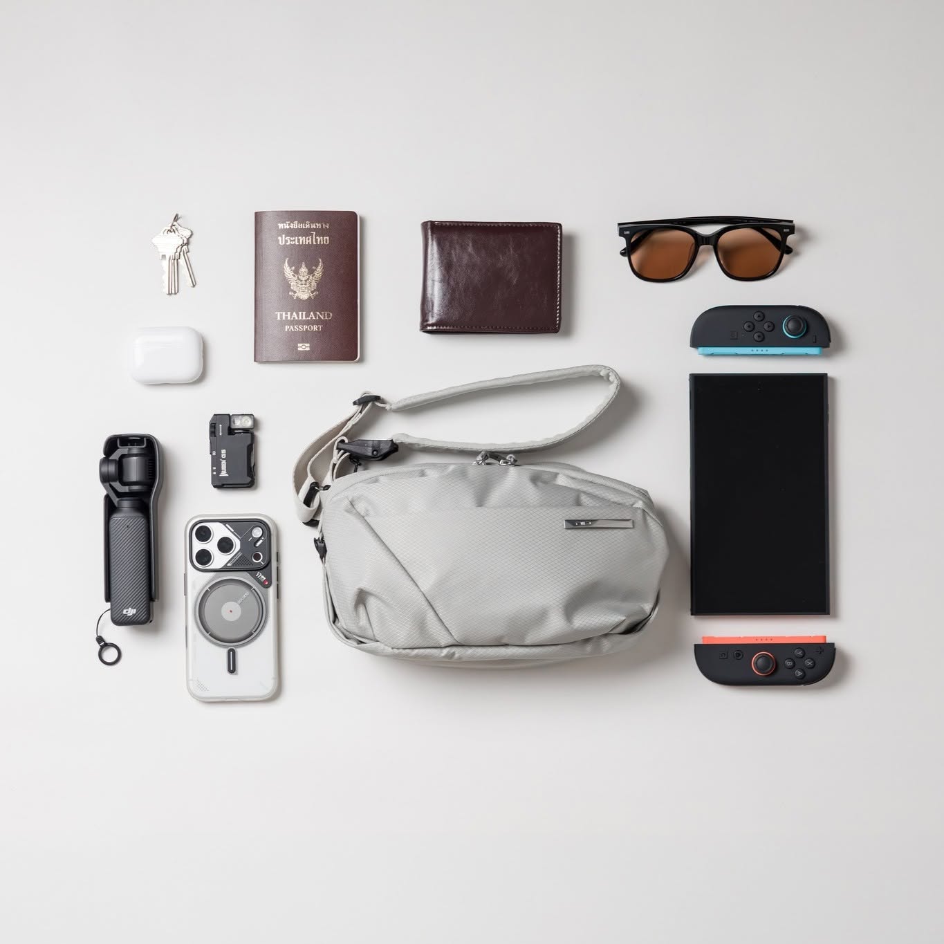 NIID NEO Nomad N1L Sling Bag in Pearly Gray shown with everyday carry items including a phone, switch console, wallet, passport, and sunglasses on a white background.