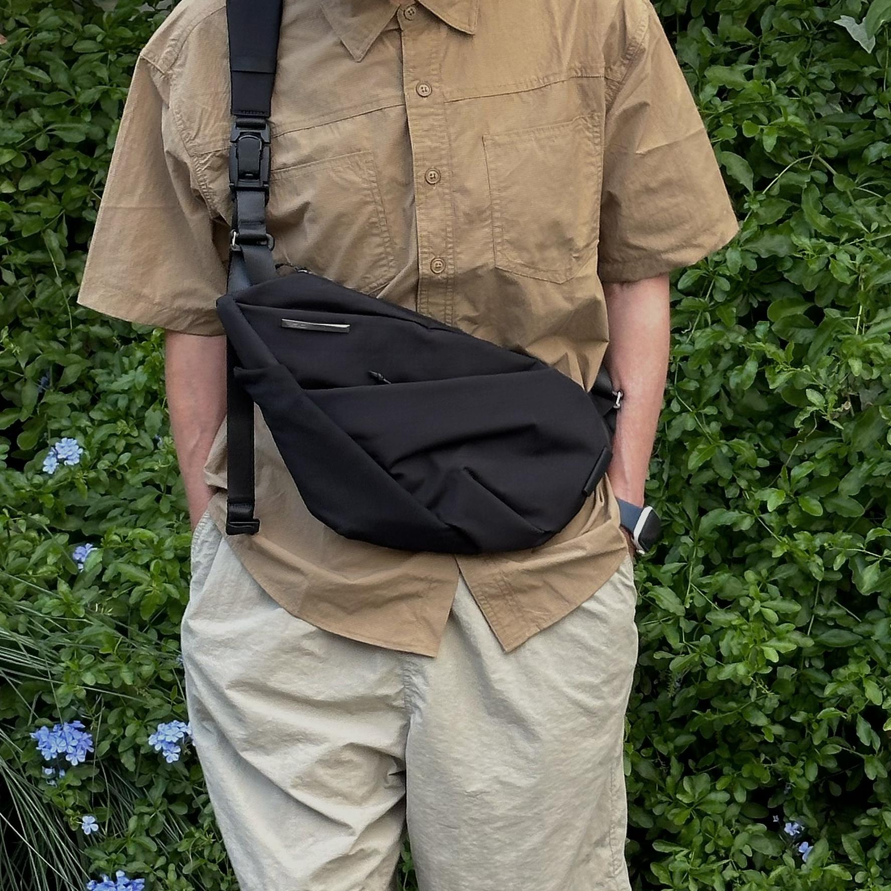 NIID FINO V Ultra‑slim Tech Sling Black Noir crossbody sling bag worn by person in tan shirt, green foliage background casual outdoor full torso shot