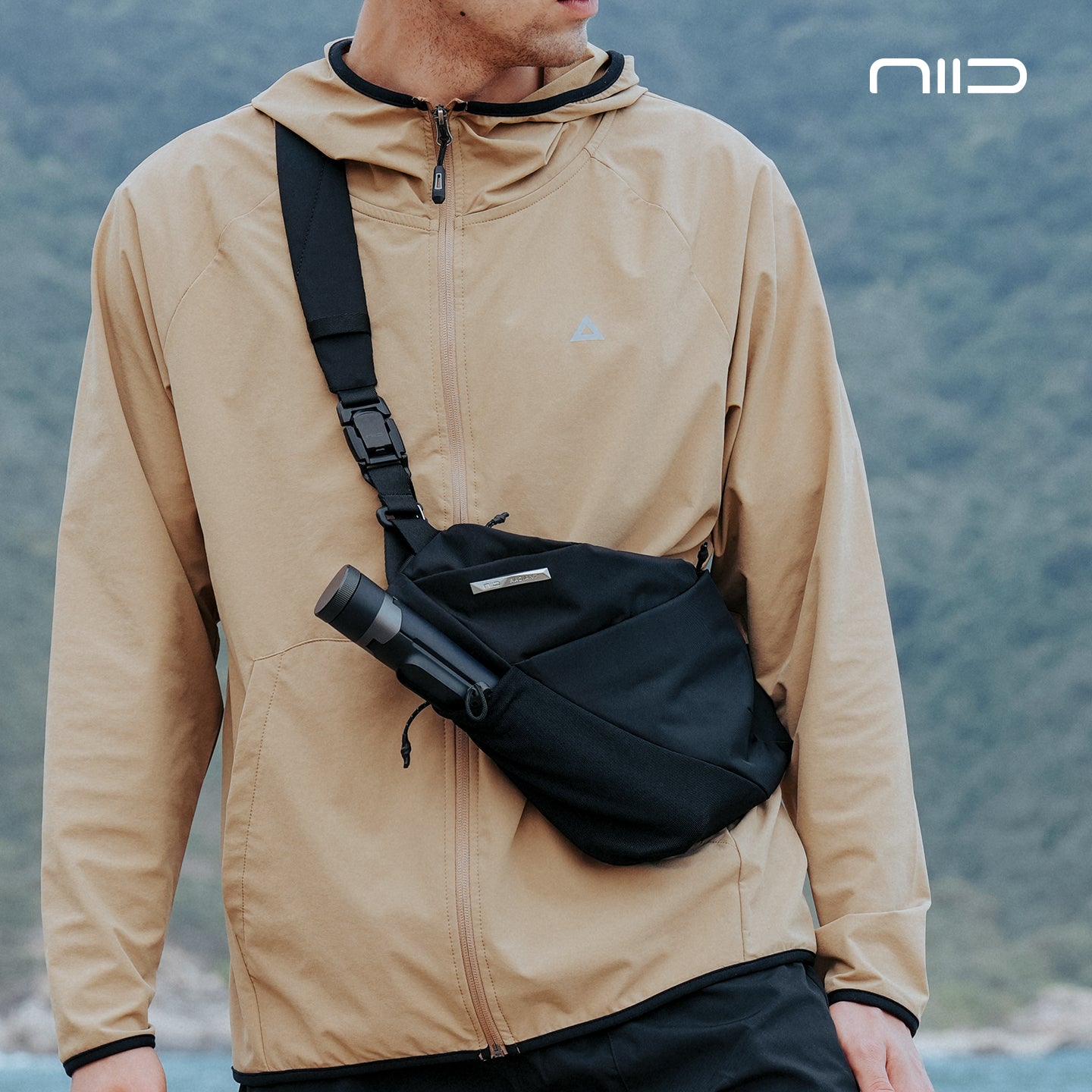 Man outdoors wearing NIID FINO V Ultra‑slim Tech Sling black noir crossbody sling bag, water bottle in front pocket, blurred mountain background