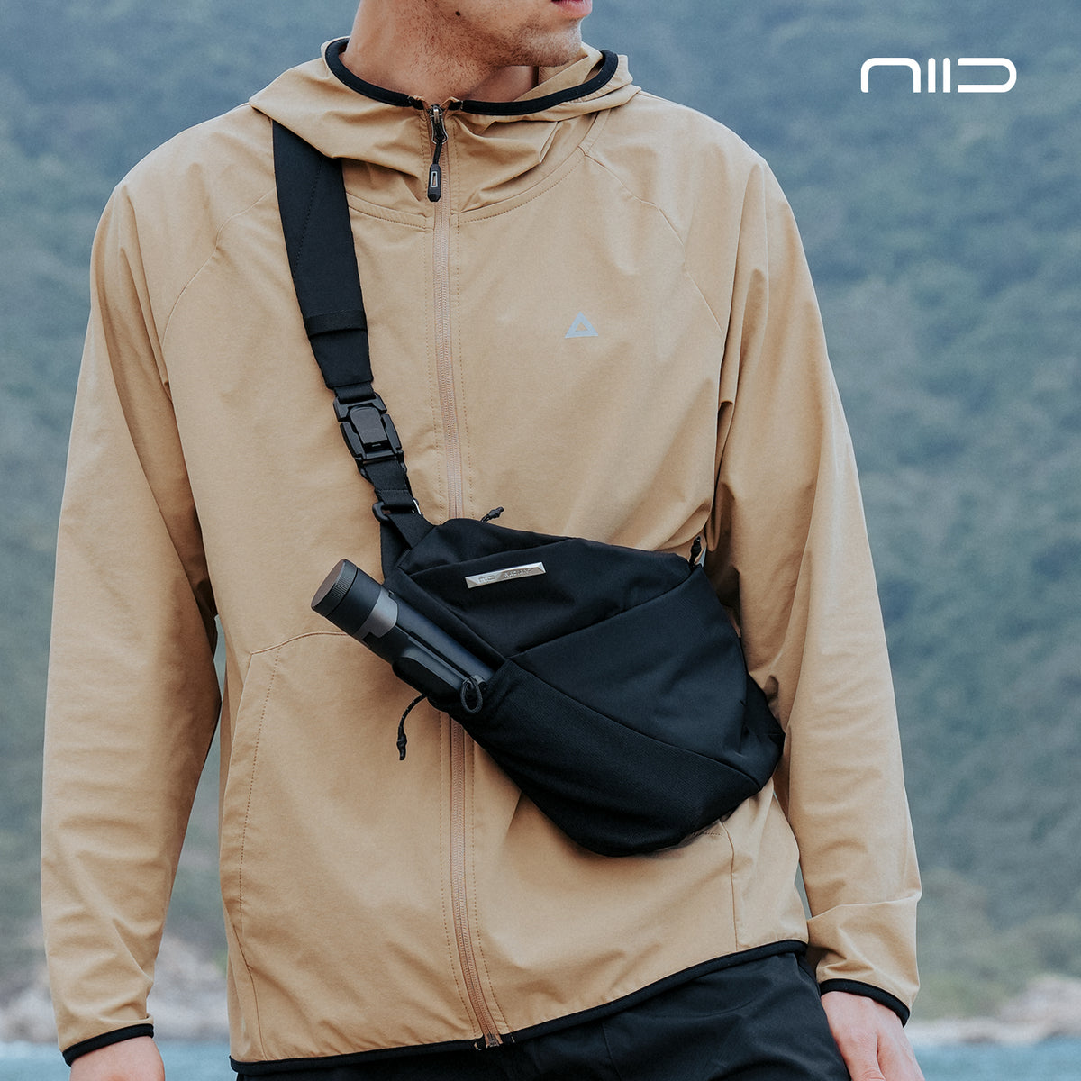 Man outdoors wearing NIID FINO V Ultra‑slim Tech Sling black noir crossbody sling bag, water bottle in front pocket, blurred mountain background