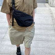 NIID FINO V Ultra‑slim Tech Sling Black Noir crossbody bag on walking person with tan shirt and khaki shorts, paved outdoor path daily‑use scene