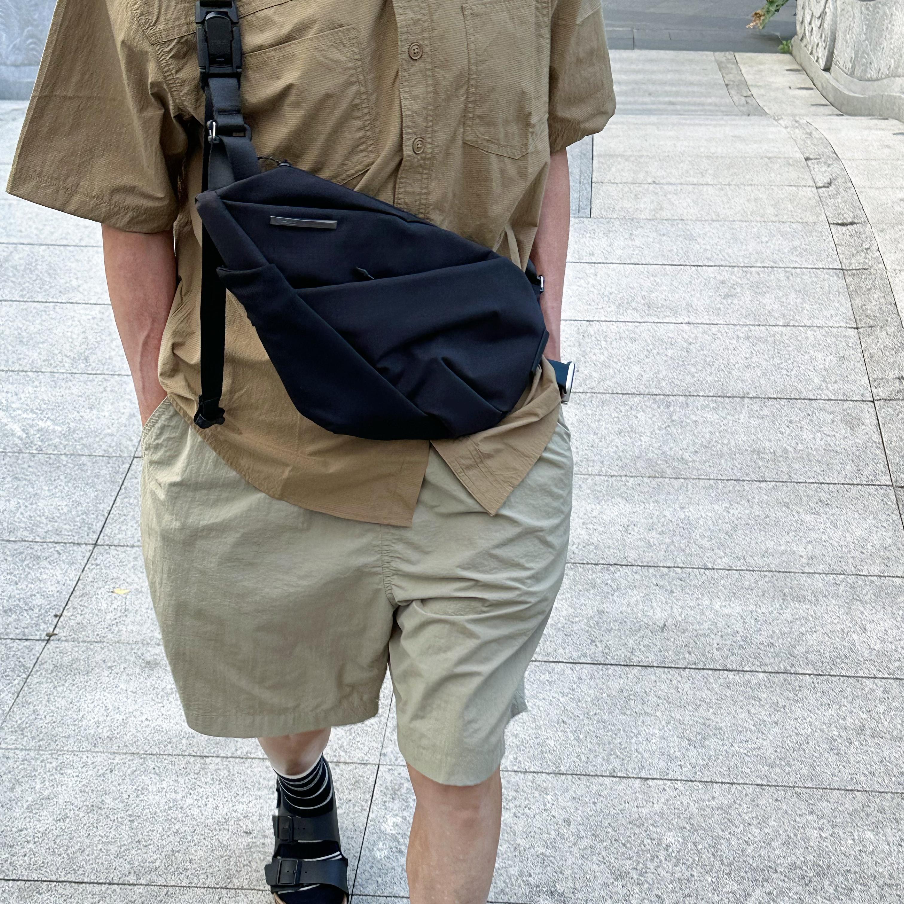 NIID FINO V Ultra‑slim Tech Sling Black Noir crossbody bag on walking person with tan shirt and khaki shorts, paved outdoor path daily‑use scene
