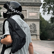 Man viewed from behind wearing NIID Radiant R2 Everyday Sling Bag, black modular crossbody sling bag with motorcycle helmet, standing before stone arch landmark in city