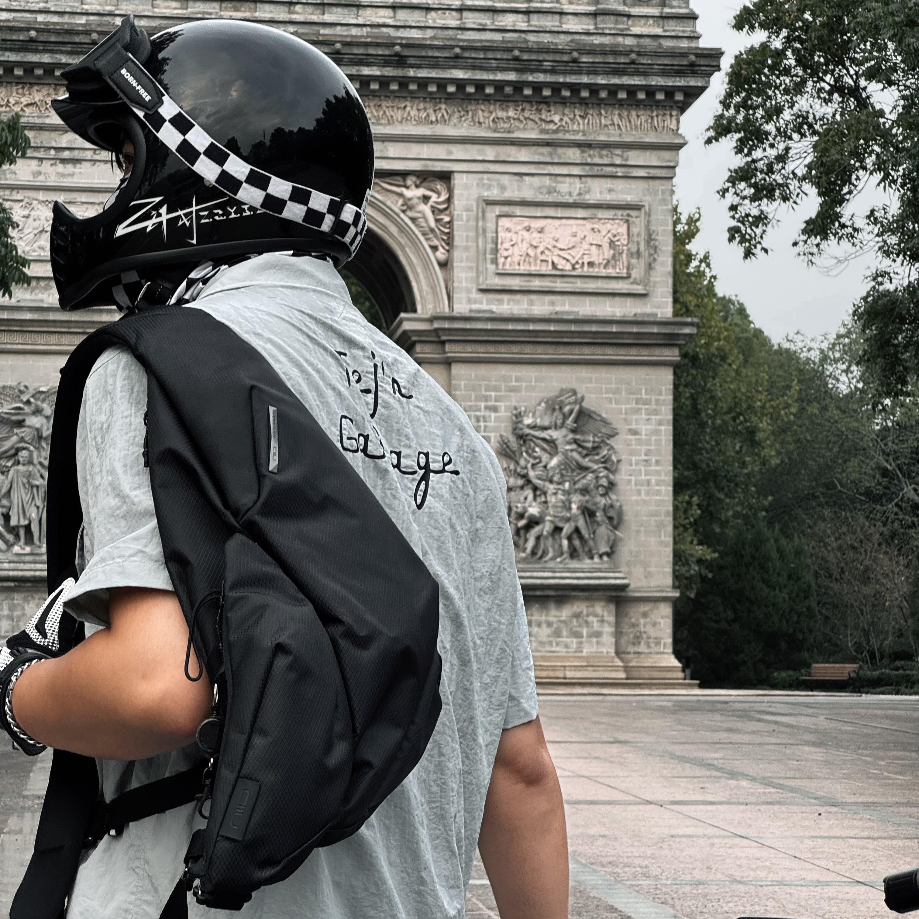 Man viewed from behind wearing NIID Radiant R2 Everyday Sling Bag, black modular crossbody sling bag with motorcycle helmet, standing before stone arch landmark in city