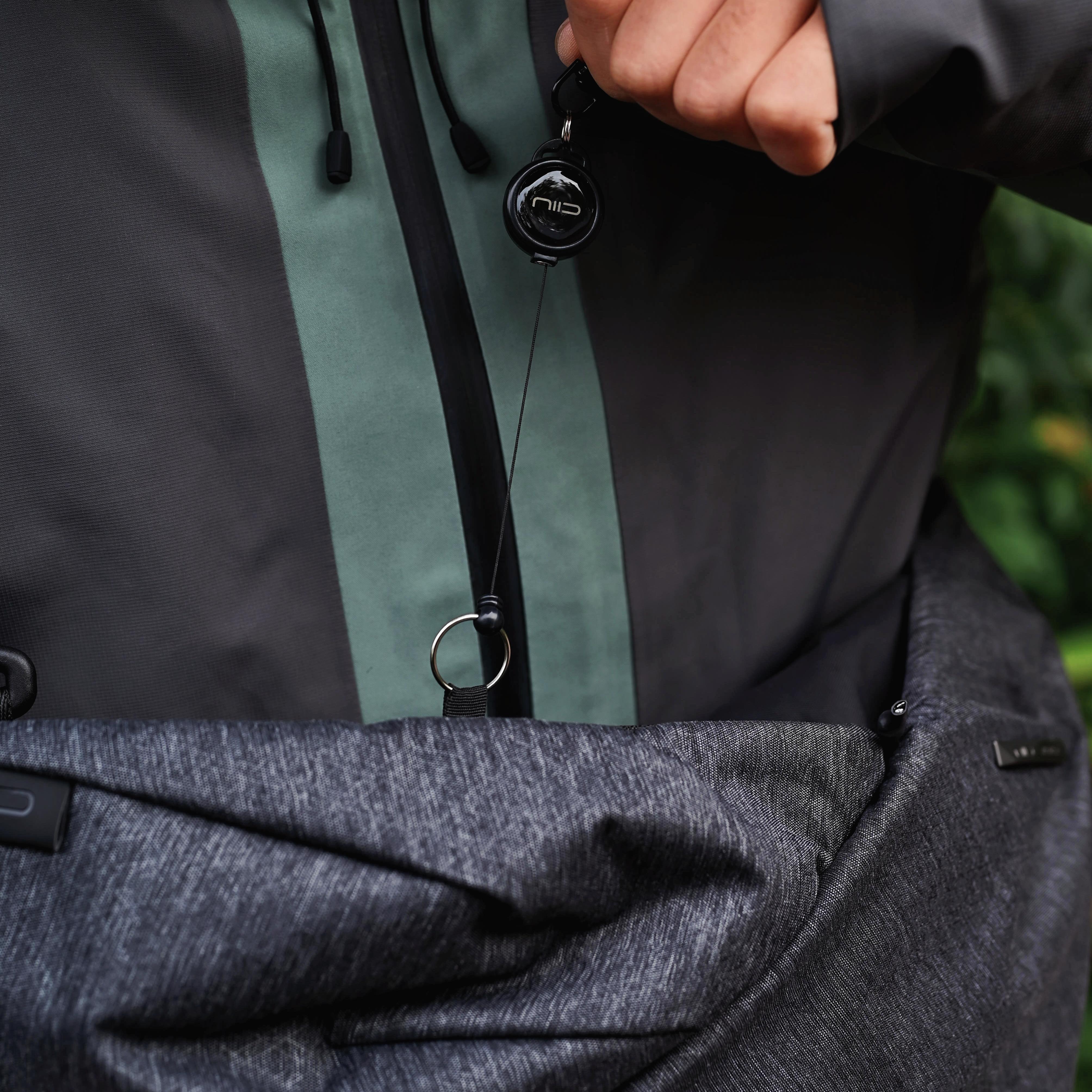 Hand holding retractable key reel accessory attached to NIID Radiant R2 Everyday Sling Bag, close‑up detail shot of bag hardware in outdoor casual setting